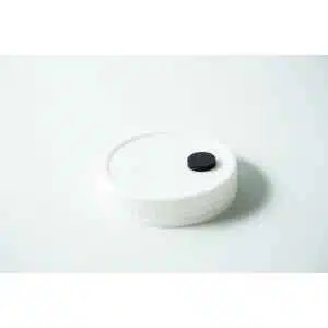 A Mycology pre-made Wide Mouth Mason Jar Lid in white with an injection port and air filter rests on a white surface. The black circular injection port is on the right, and the textured white air filter is on the left of the lid.