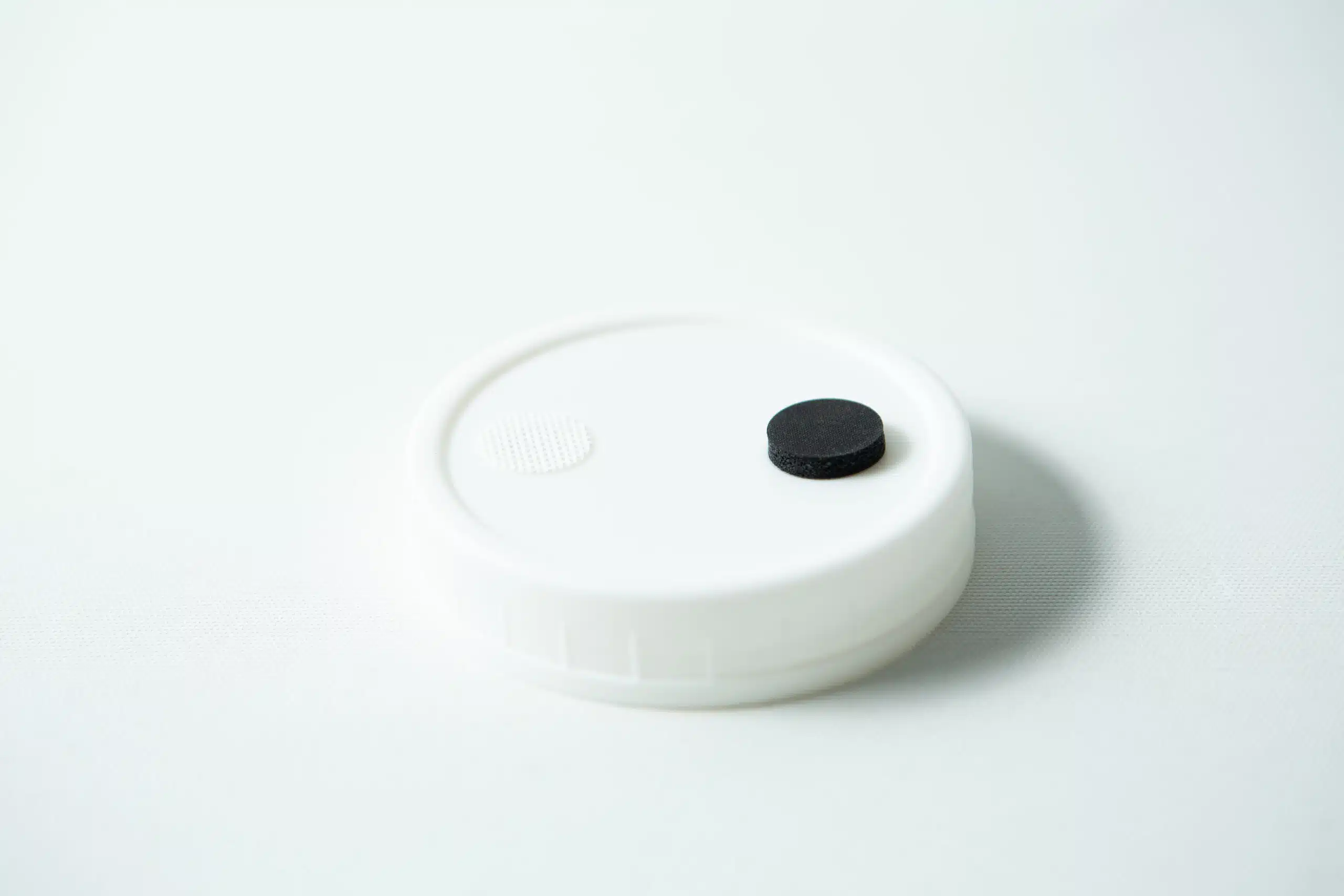 Mycology premade Wide Mouth Mason Jar Lids white with inj port and air filter 1 A Mycology pre-made Wide Mouth Mason Jar Lid in white with an injection port and air filter rests on a white surface. The black circular injection port is on the right, and the textured white air filter is on the left of the lid.