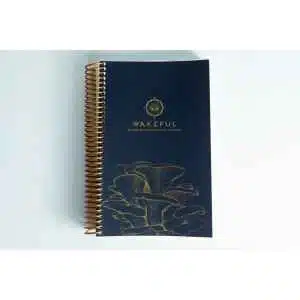 A navy blue spiral-bound Wakeful Mushroom Cultivation Journal by Australian mycologist Caine Barlow, featuring a gold mushroom line drawing on the cover, rests closed on a white surface.