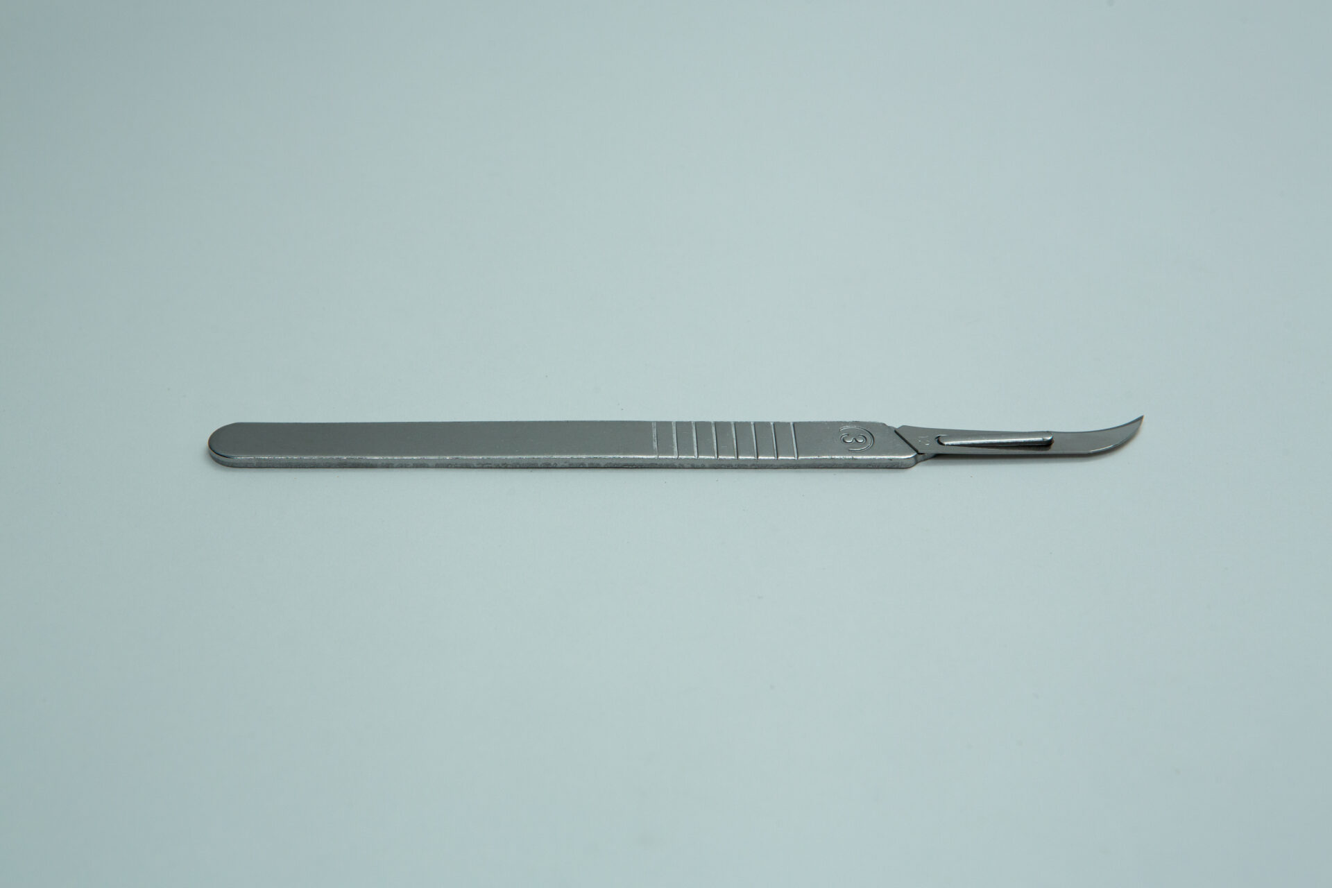Scalpel handle #3 and blades | Mycology 8 A single scalpel handle no. 3 with a curved blade for mycology is placed horizontally on a plain light grey background, highlighting the precision of its finely honed metal blade.