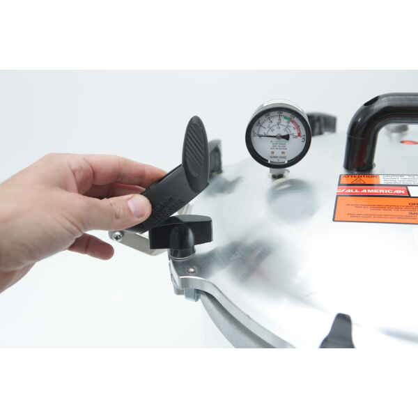 A hand lifts the locking lever of an All American Steriliser lid using the Rootlab Original 3D Printed Wingnut Spanner, with a pressure gauge and black handle visible on top.