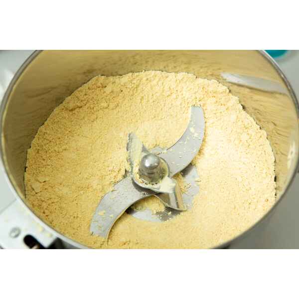 Close-up of a metal food processor bowl containing finely ground yellow powder with metal blades in the centre.