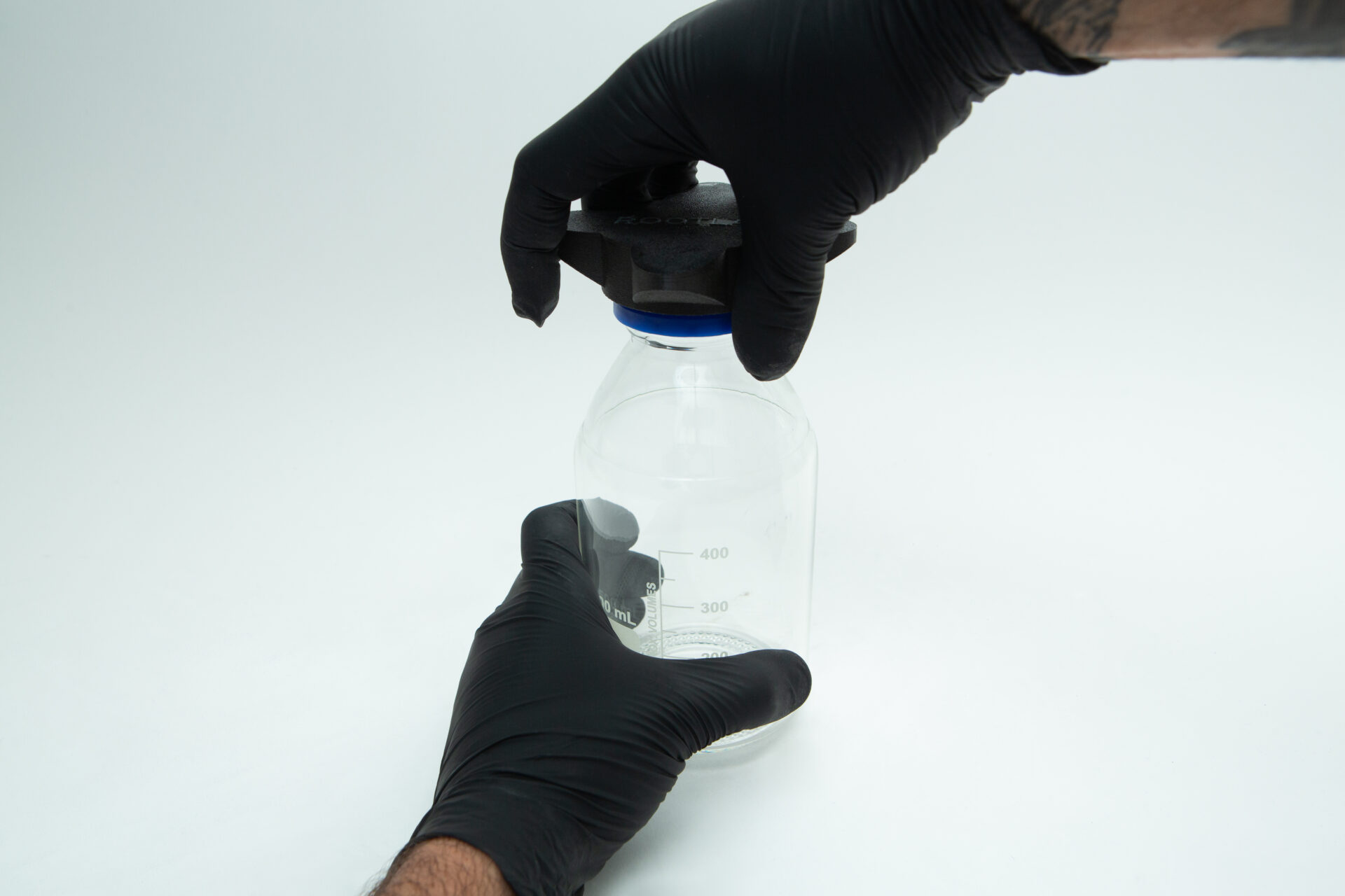 Reagent Bottle Cap Wrench | Rootlab Original | 3D Printed 5 A person wearing black gloves is placing or removing a black cap on a clear glass laboratory bottle with measurement markings, against a plain white background.