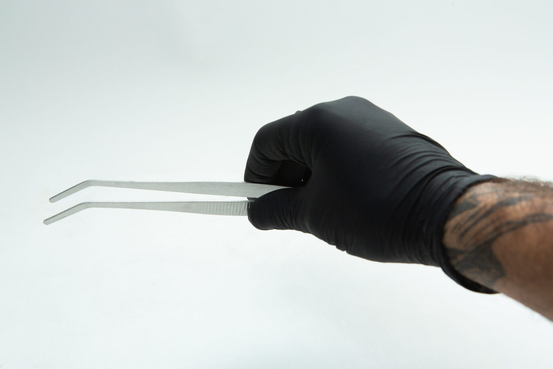 Stainless Steel Plant tissue culture Lab Forceps | Curved & Straight Tips | 25cm 4 A hand wearing a black glove holds a pair of angled metal tweezers against a plain white background. A tattoo is visible on the person's forearm.
