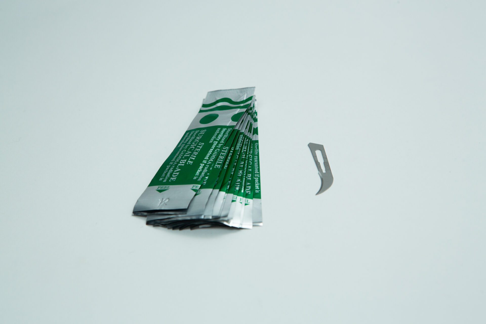 Scalpel handle #3 and blades | Mycology 5 A stack of individually wrapped green and silver razor blades is placed on a white surface, with one unwrapped curved razor blade lying next to the stack.