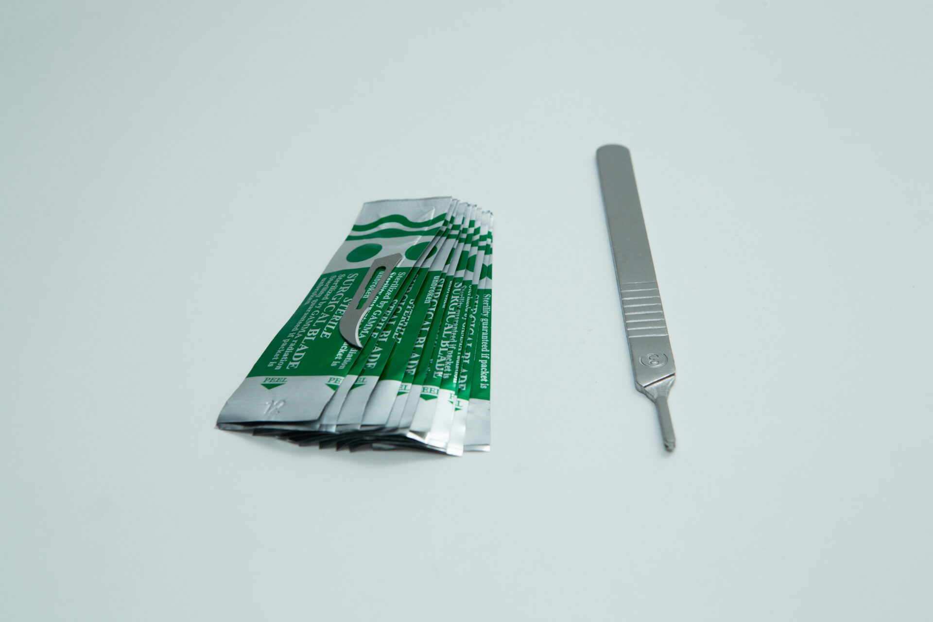 Scalpel handle #3 and blades | Mycology 6 A metal scalpel handle with a blade attached is placed next to several individually wrapped scalpel blades with green and silver packaging, displayed on a plain white background.