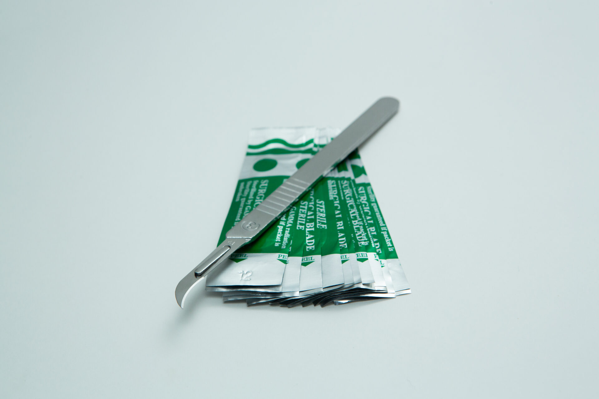 Scalpel handle #3 and blades | Mycology 7 A metal scalpel with a curved blade rests on top of several green and silver sterile blade packets, arranged on a plain white background.