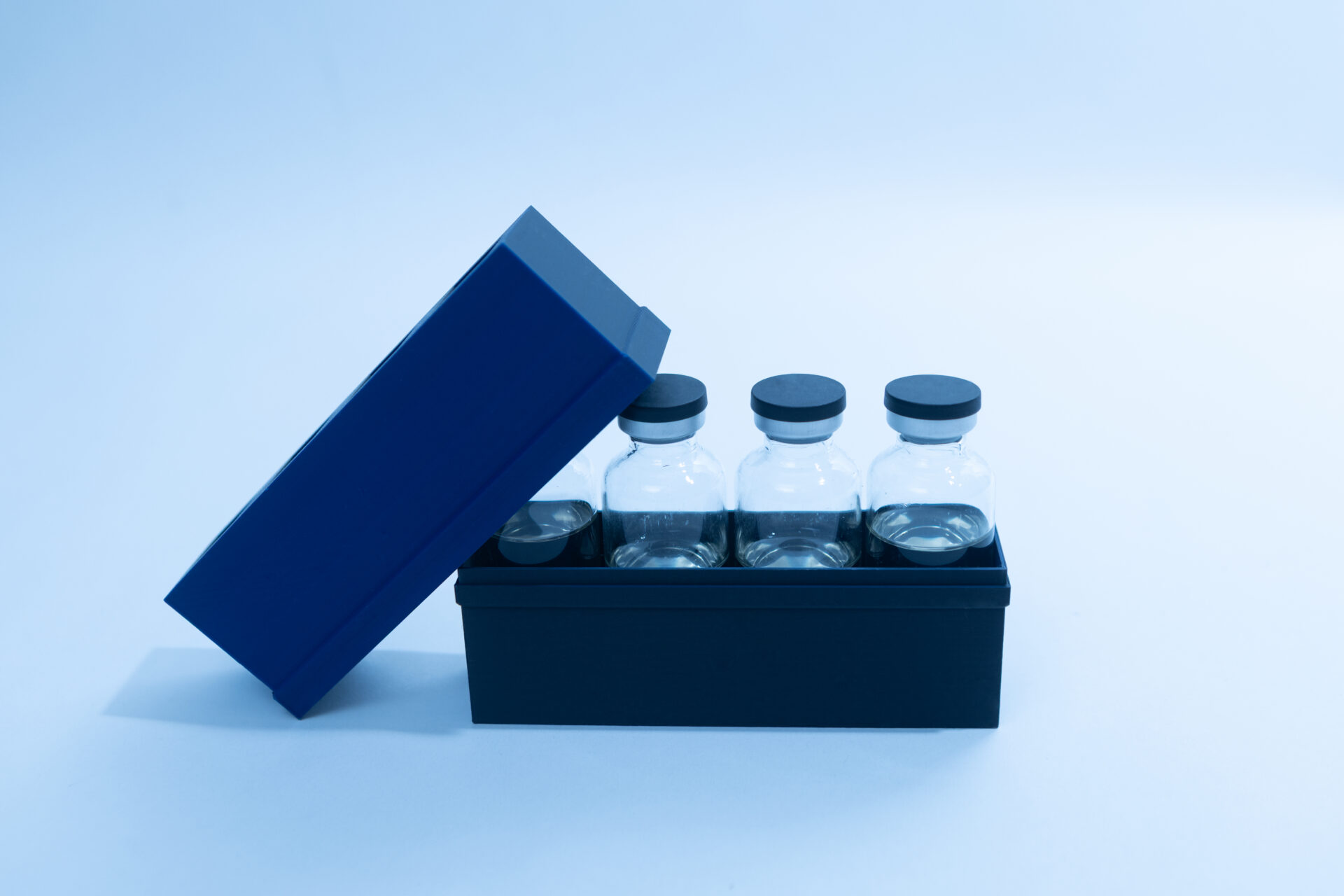 Sterile Liquid Culture Vials, Dr Myc Adaptive LC, Crimp Sealed 28 A black box with four empty glass vials, each sealed with a black cap, sits on a light blue surface. The box lid is partially open and tilted. The background and lighting are blue-toned.