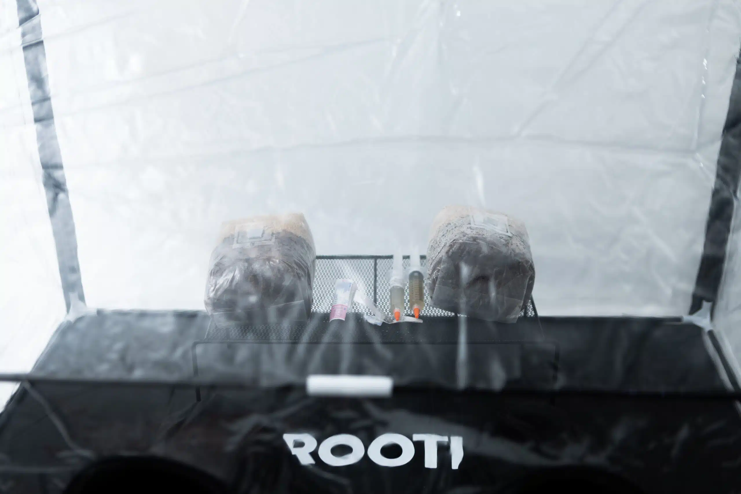 Rootlab Still Air Box with angled viewing window- XL 4 Two seats wrapped in plastic are visible behind a mesh pocket containing various bottles and items, all inside a transparent enclosure. The partial word ROOTI is printed on the front of a black surface.