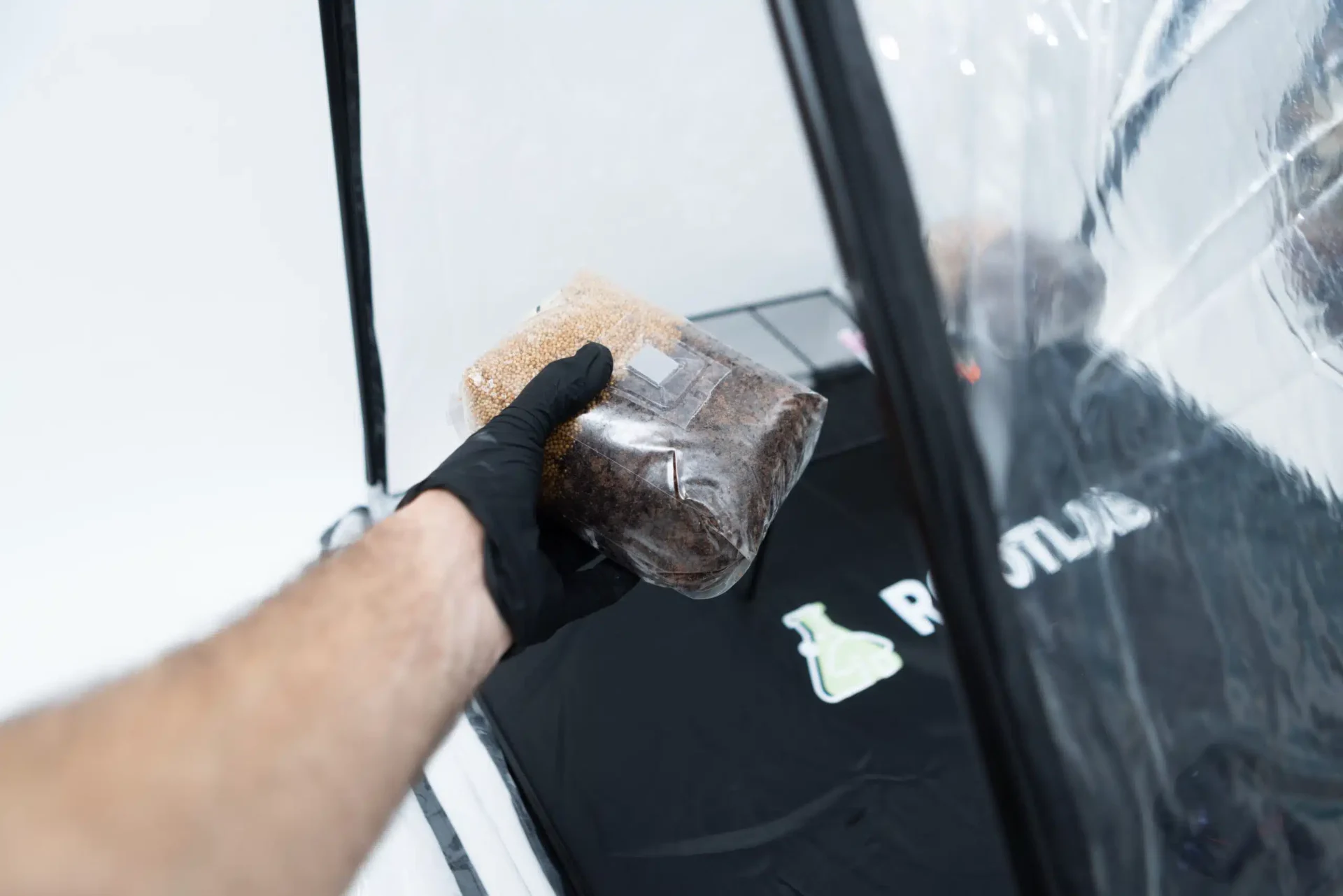 Rootlab Still Air Box with angled viewing window- XL 5 A person wearing a black glove holds a sealed plastic bag of brown and beige substrate inside a clear, tent-like enclosure with a black mat labelled RUSTIC..