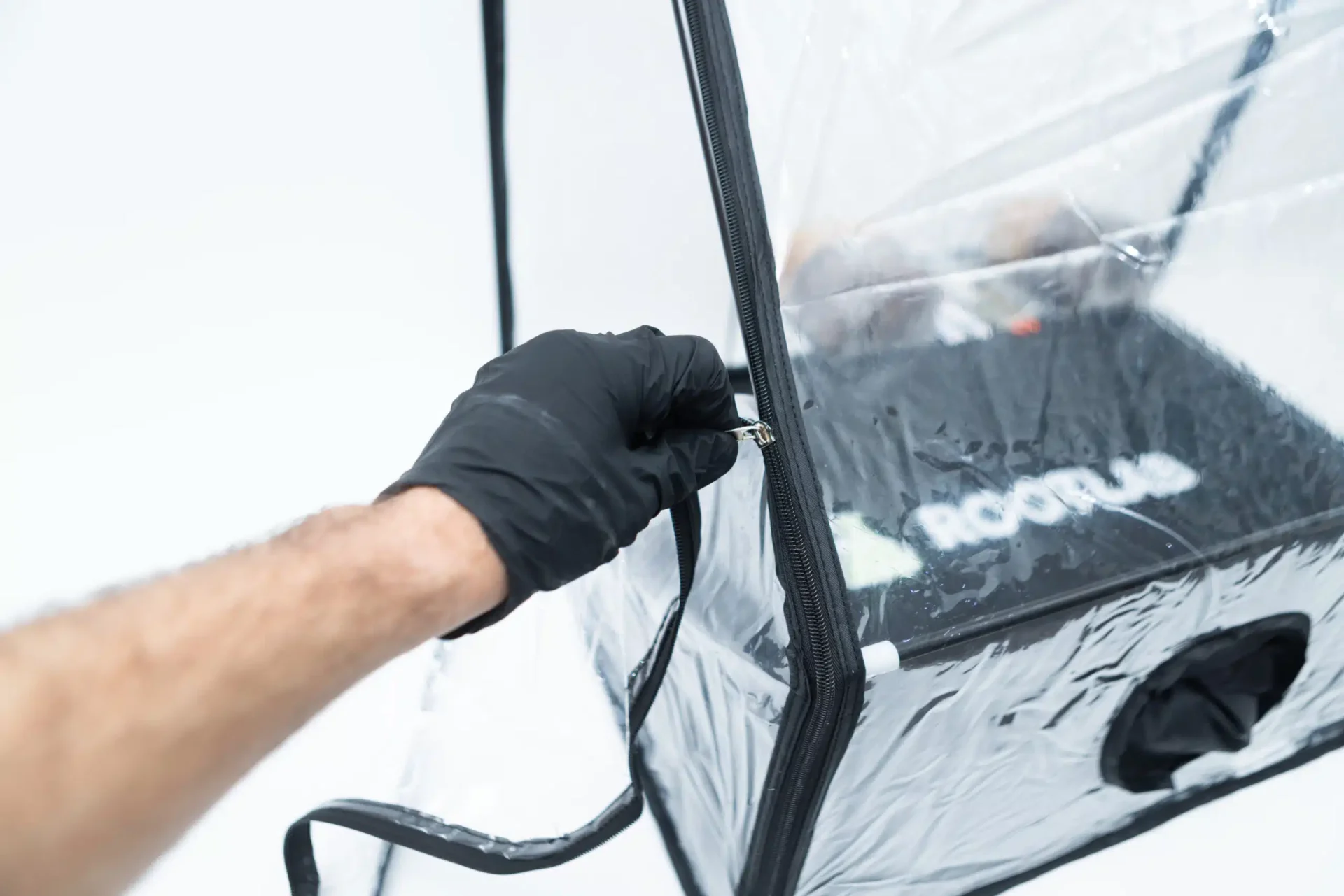 Rootlab Still Air Box with angled viewing window- XL 6 A person wearing a black glove is unzipping a transparent plastic enclosure. Inside the enclosure, a black object with the word ROOM partially visible is placed. The background is plain white.