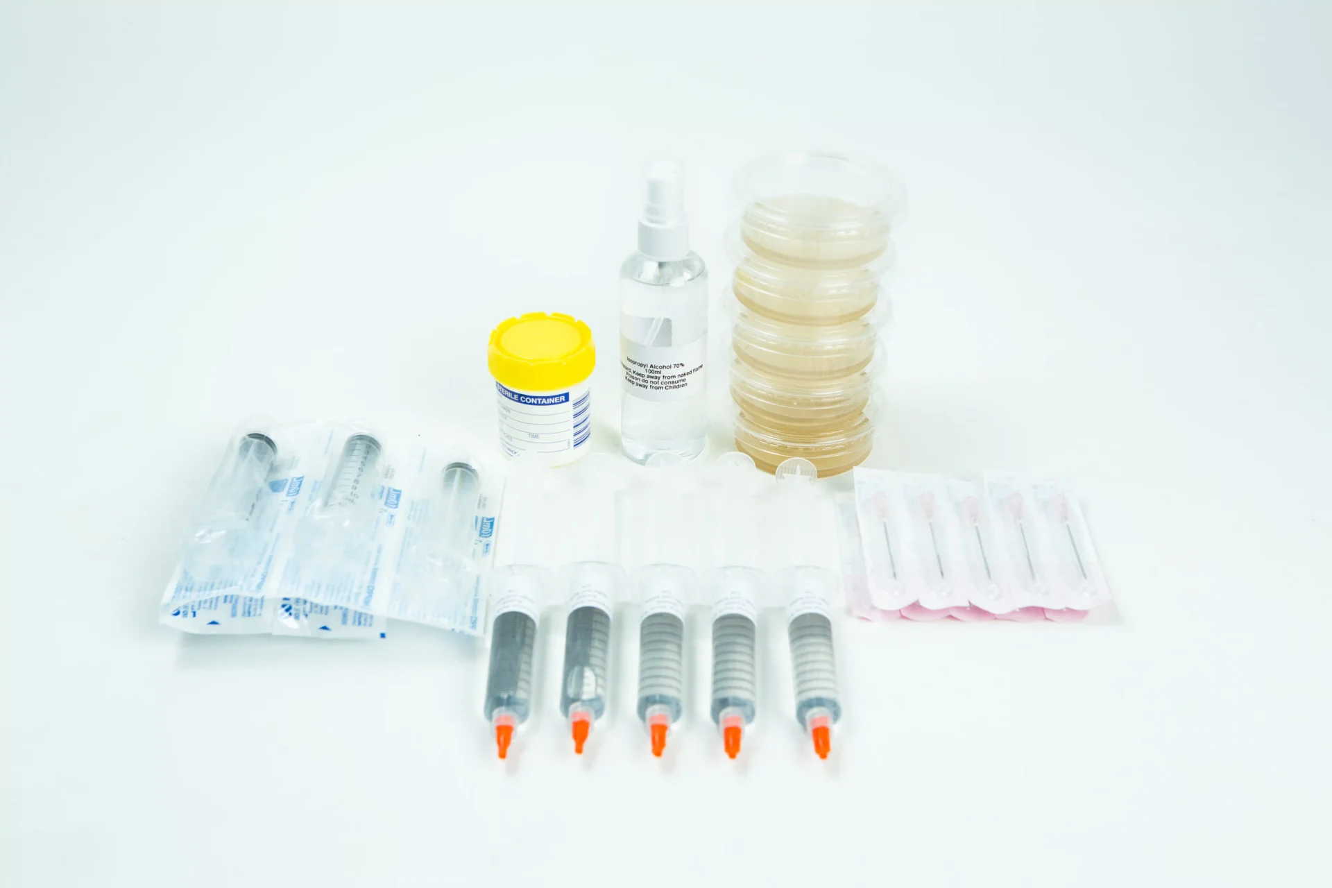 Spore Print to Spore syringe transfer Kit 1 Several sealed medical syringes, a yellow-capped specimen container, a clear spray bottle, stacked Petri dishes, and packaged items are arranged on a white surface.