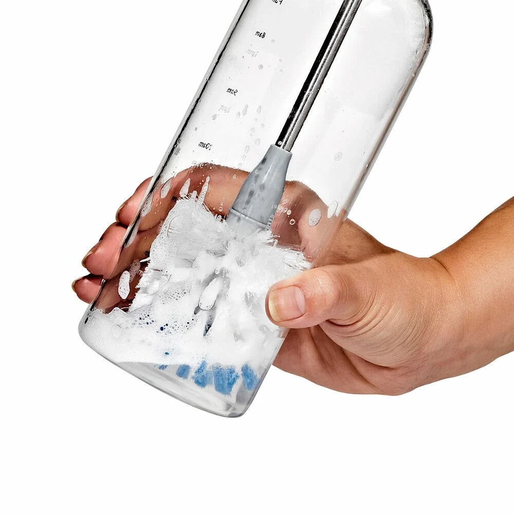 OXO Good Grip Stainless Steel Bottle Brush with replaceable head 4 A hand holds a clear bottle whilst using a brush to clean the inside. The bottle has measurement markings, and soapy water with bubbles is visible inside.