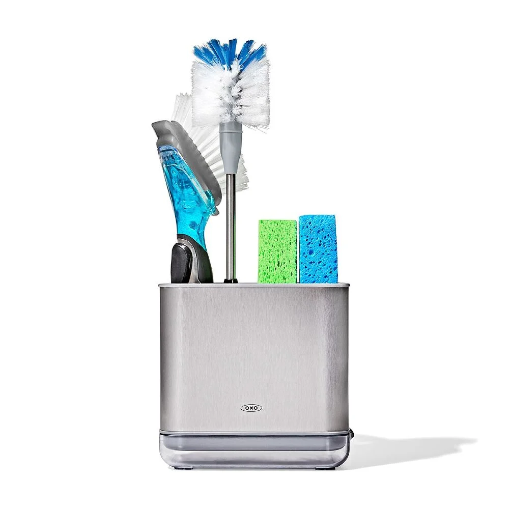 OXO Good Grip Stainless Steel Bottle Brush with replaceable head 5 A stainless steel kitchen caddy holds a scrubbing brush, a washing-up brush with a blue handle, and two rectangular sponges—one green and one blue—against a white background.