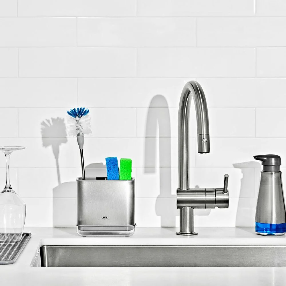 OXO Good Grip Stainless Steel Bottle Brush with replaceable head 6 A kitchen sink with a modern tap, a silver organiser holding a brush and sponges, a soap dispenser, a glass, and a drying rack, set against a white tile splashback.